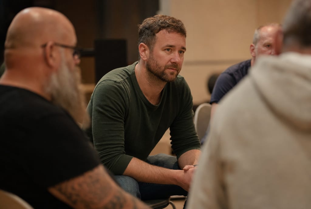A man in a support-group circle leaning forward and listening, tired but resolved.